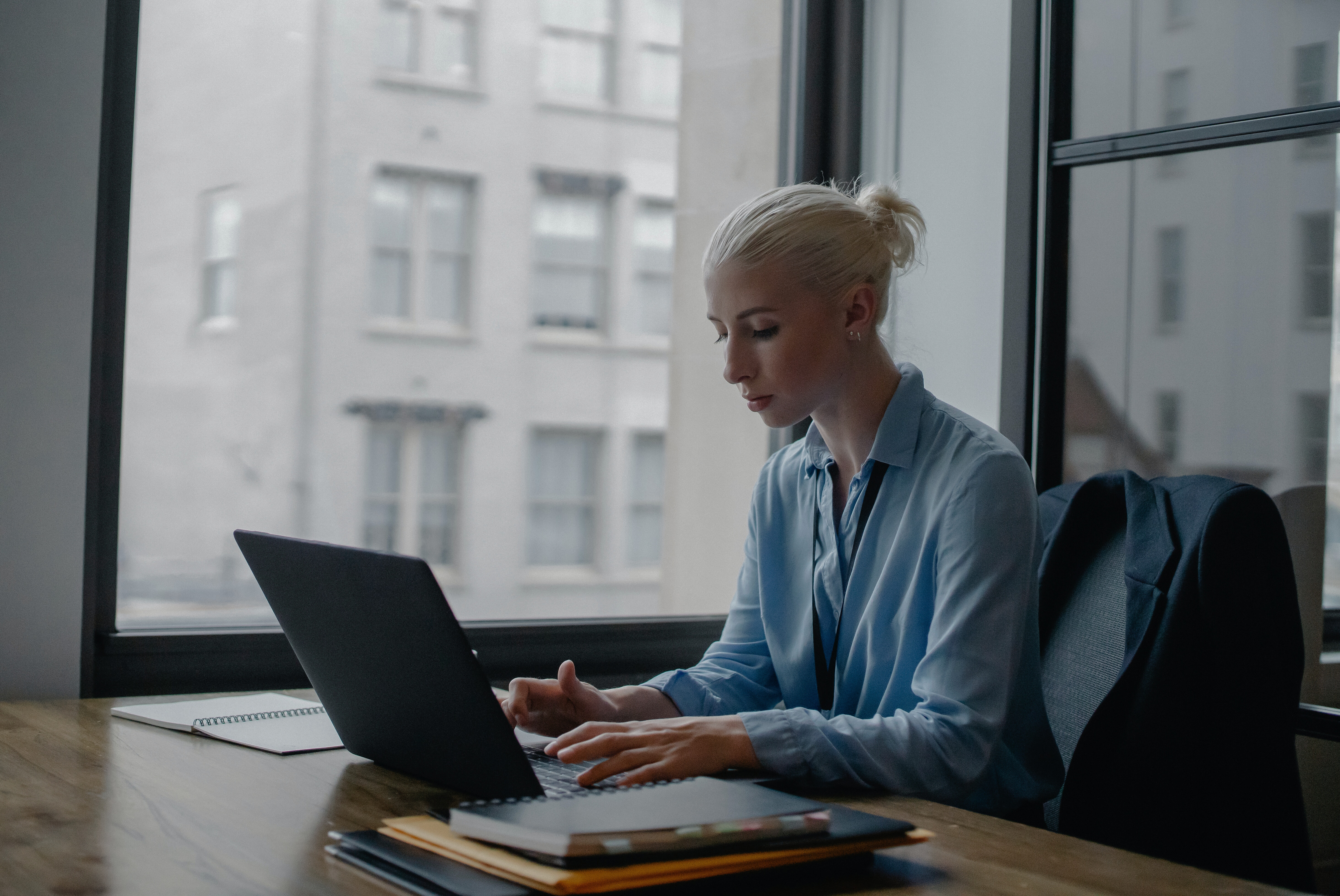 A young professional woman works on a laptop next to a large window overlooking a skyscraper.